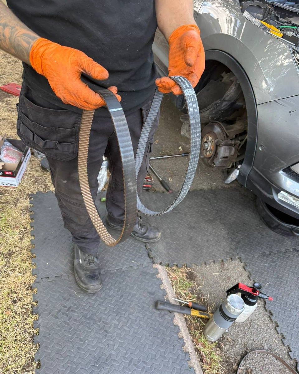 The EH Autos mechanic holding a worn timing belt beside its replacement at a job